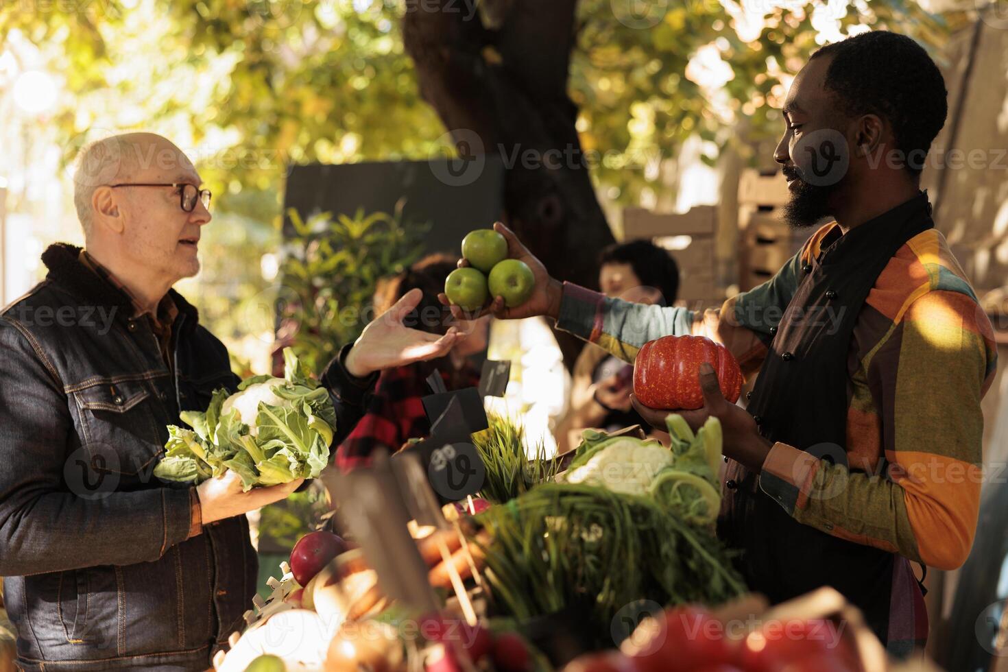 Senior man shopping for local organic produce at farmers market on weekend. Young African guy vendor selling locally grown natural fruits and vegetables to satisfied elderly male customer photo