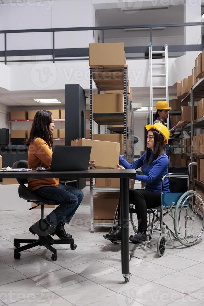 Asian postal service worker in wheelchair and operator maintaining parcel delivery. Storehouse order packer with disability giving cardboard box to distribution manager in storage room photo
