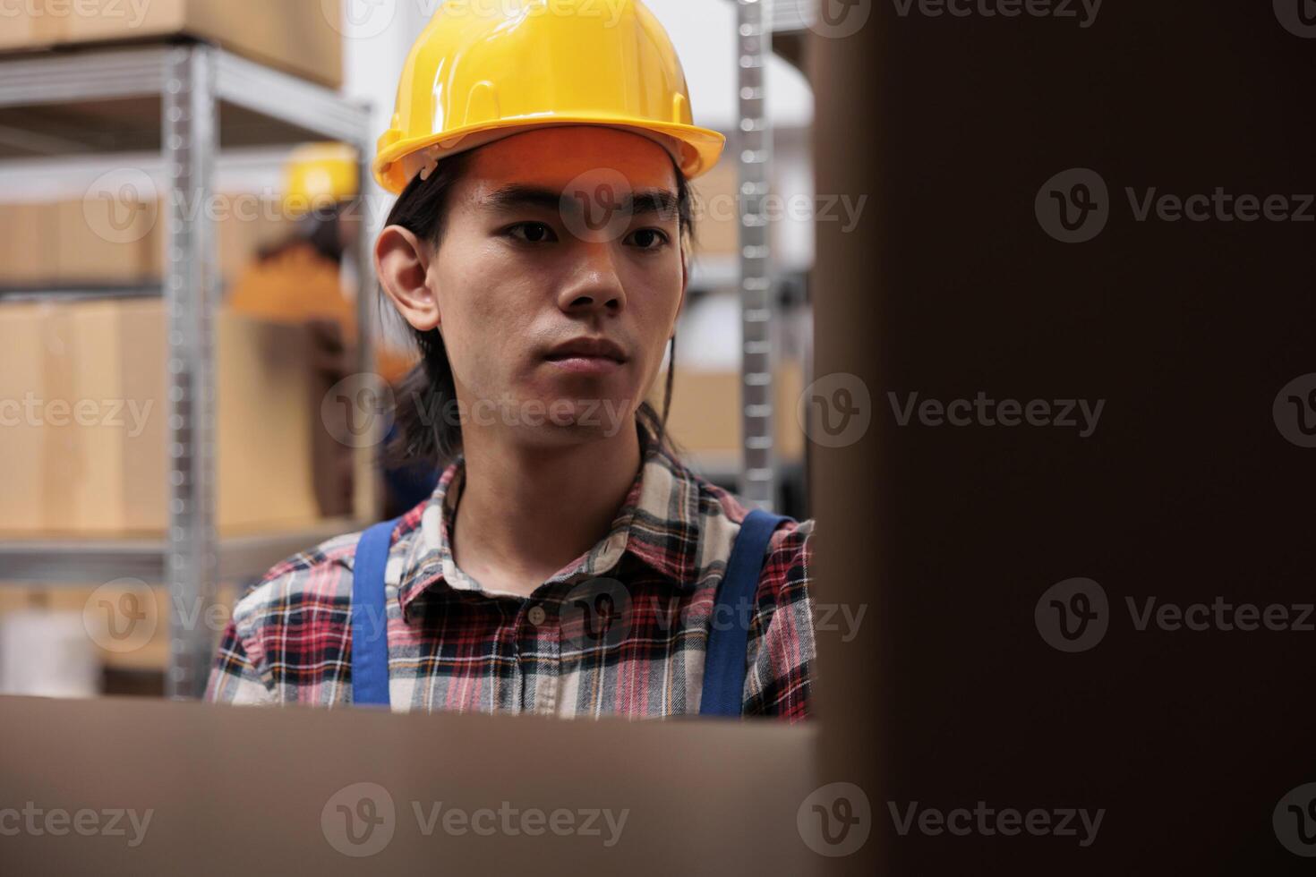 Warehouse employee in yellow helmet searching parcel on shelf while working in storage room. Young asian delivery service worker managing cardboard boxes storing in storehouse photo