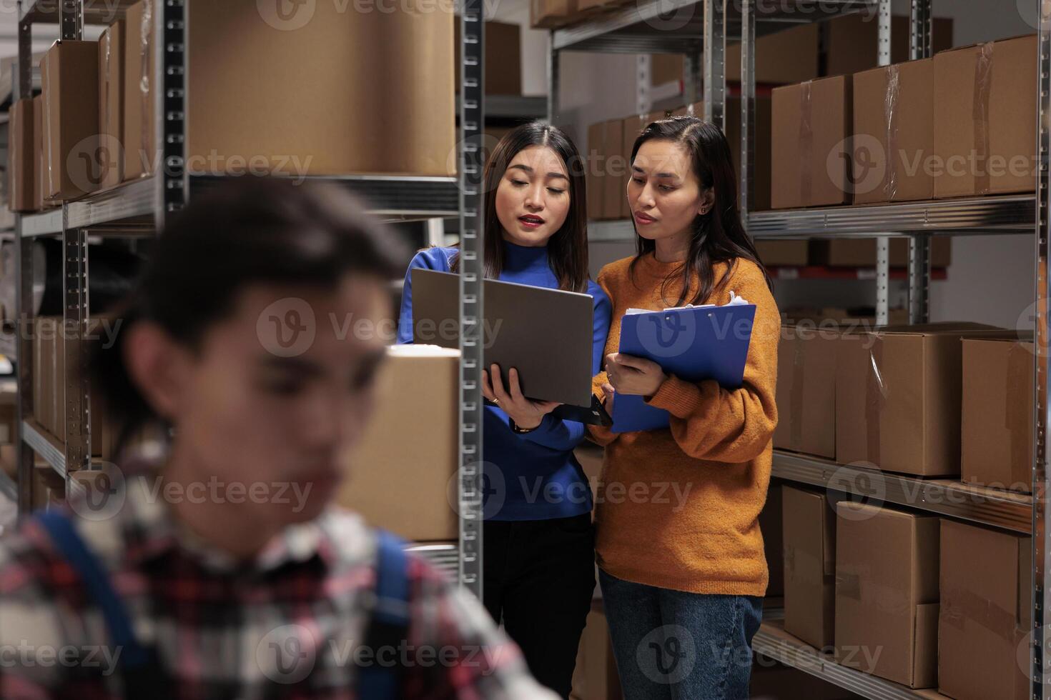 Warehouse supervisor teaching trainee managing inventory on laptop. Asian storehouse manager showing freight distribution report to woman coworker in storage room full of cardboard boxes photo