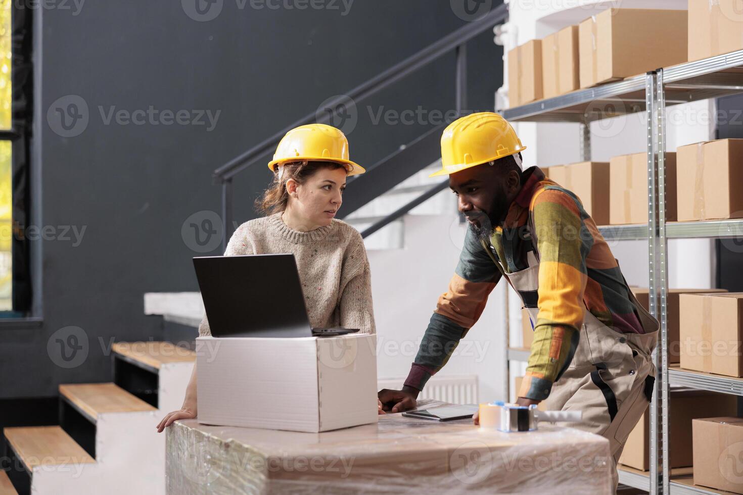 Warehouse team using boxes to pack customers orders, analyzing delivery details on laptop computer in storage room. African american employee wearing industrial overall and helmet during inventory photo