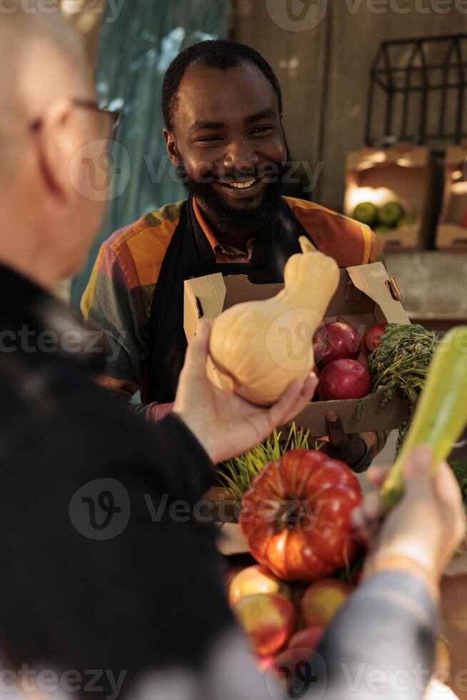 Fresh products for elderly client at organic food market, man talking to vendor and shopping at local farmers market. Friendly farmer standing behind fruit and vegetable stall and serving customer. photo