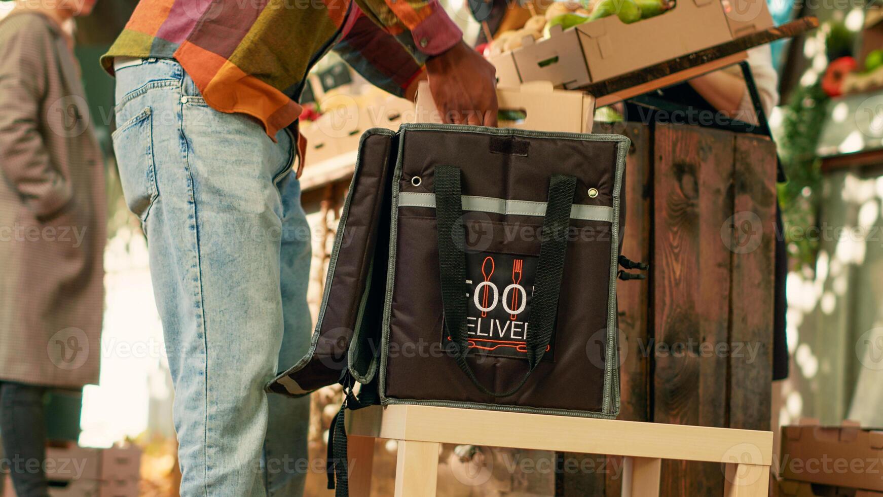 Young happy courier picking up bio products order from local farmers market, delivering healthy homegrown food to clients at home. Deliveryman working with team of farmers at stand. Handheld shot. photo