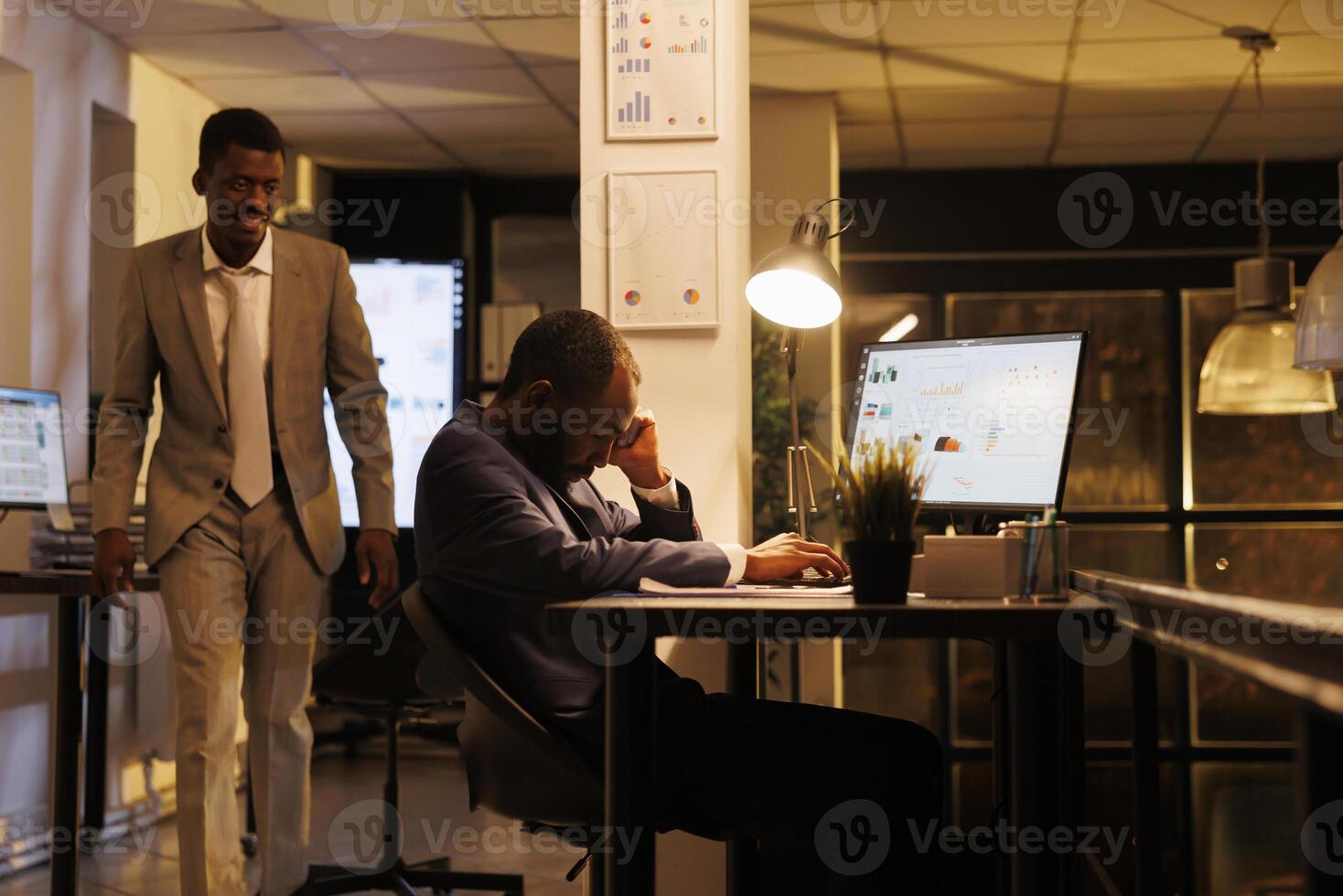Manager wakes up employee after falling asleep on table desk in startup office. African american colleagues working overhours, planning financial investment strategy to increase company profit photo