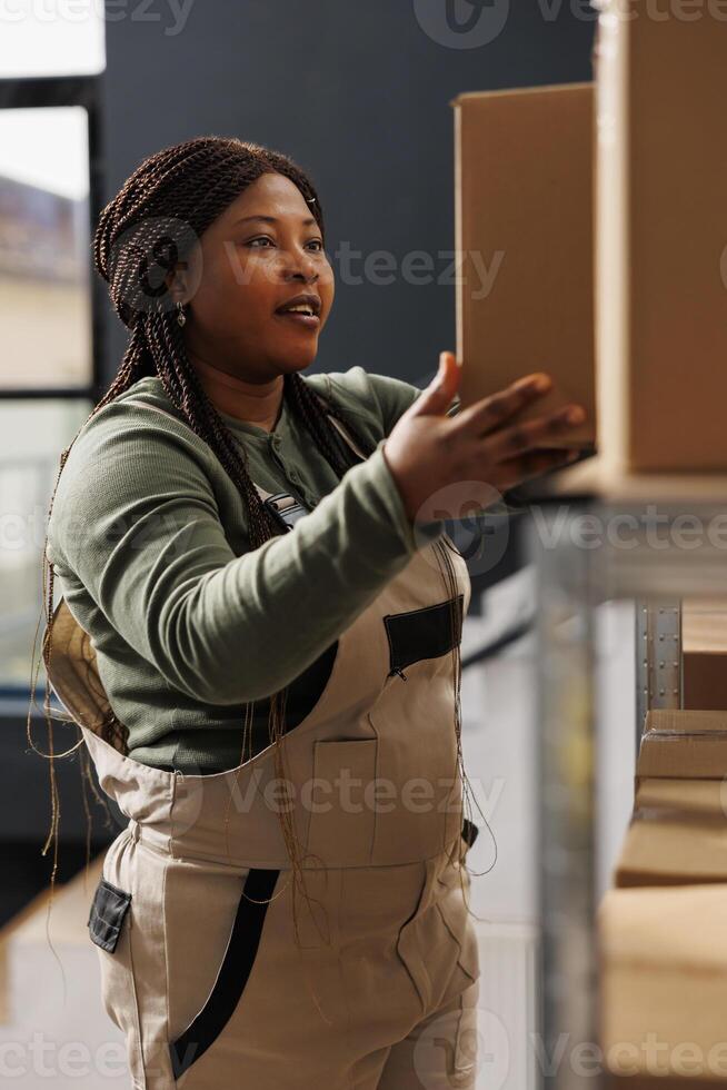 Cheerful manager taking out cardboard box from shelves, preparing customers order during work shift in warehouse. African american employee wearing industrial overall working at products delivery photo