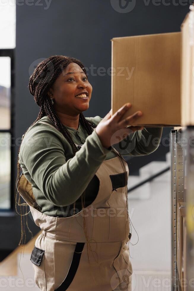 Stockroom worker taking out cardboard box from shelves, preparing customers online orders in warehouse. African american supervisor wearing industrial overall working at products delivery in storehouse photo