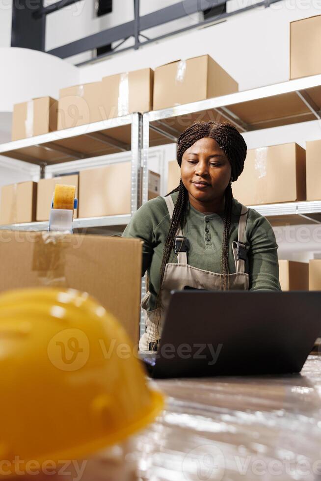 African american supervisor using cardboard boxes to pack customers orders, analyzing shipping details on laptop computer in storage room. Warehouse worker wearing industrial overall during inventory photo