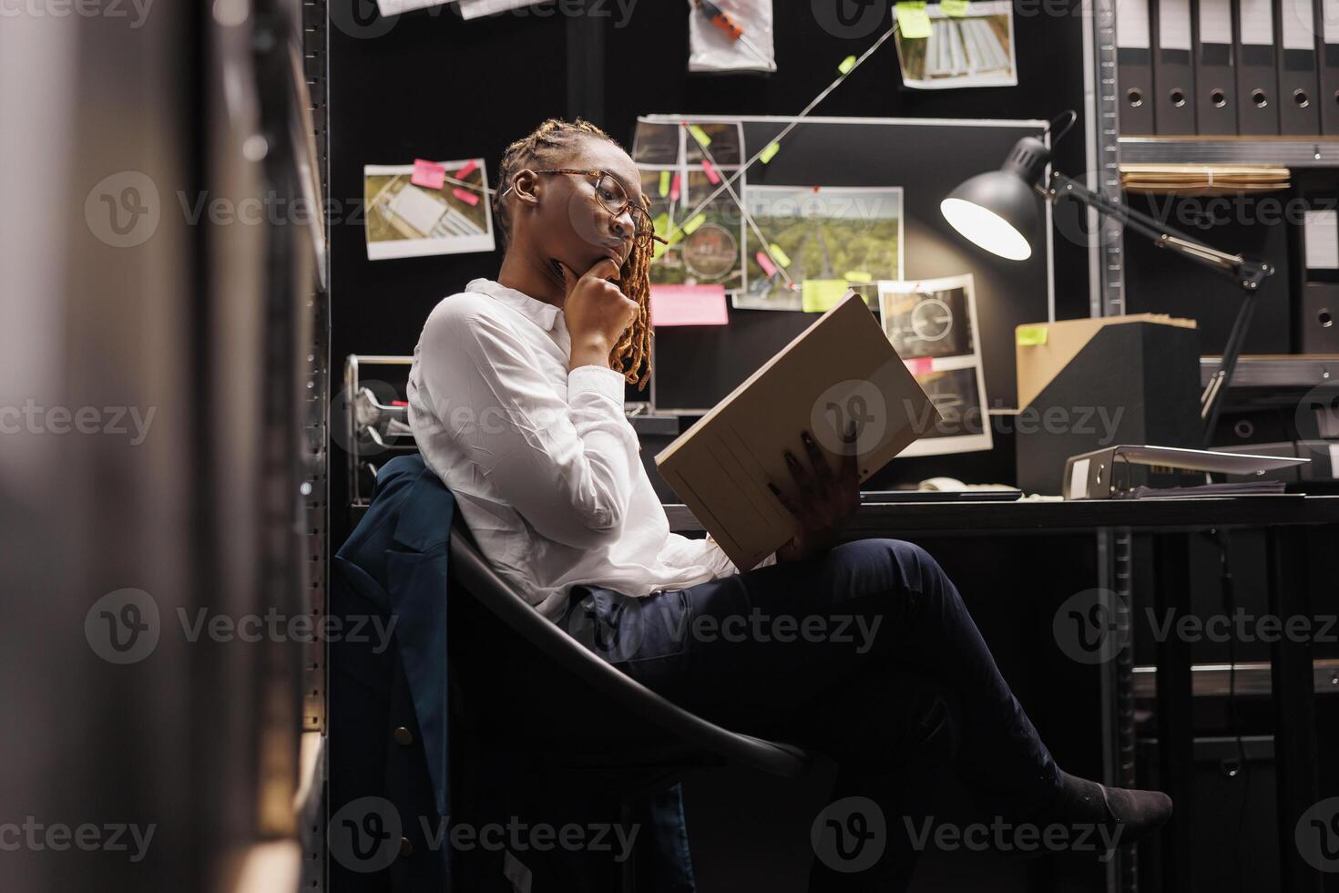 Thoughtful woman investigator reviewing forensic expertise report and rubbing chin in police office room. African american detective reading crime case file and analyzing information photo