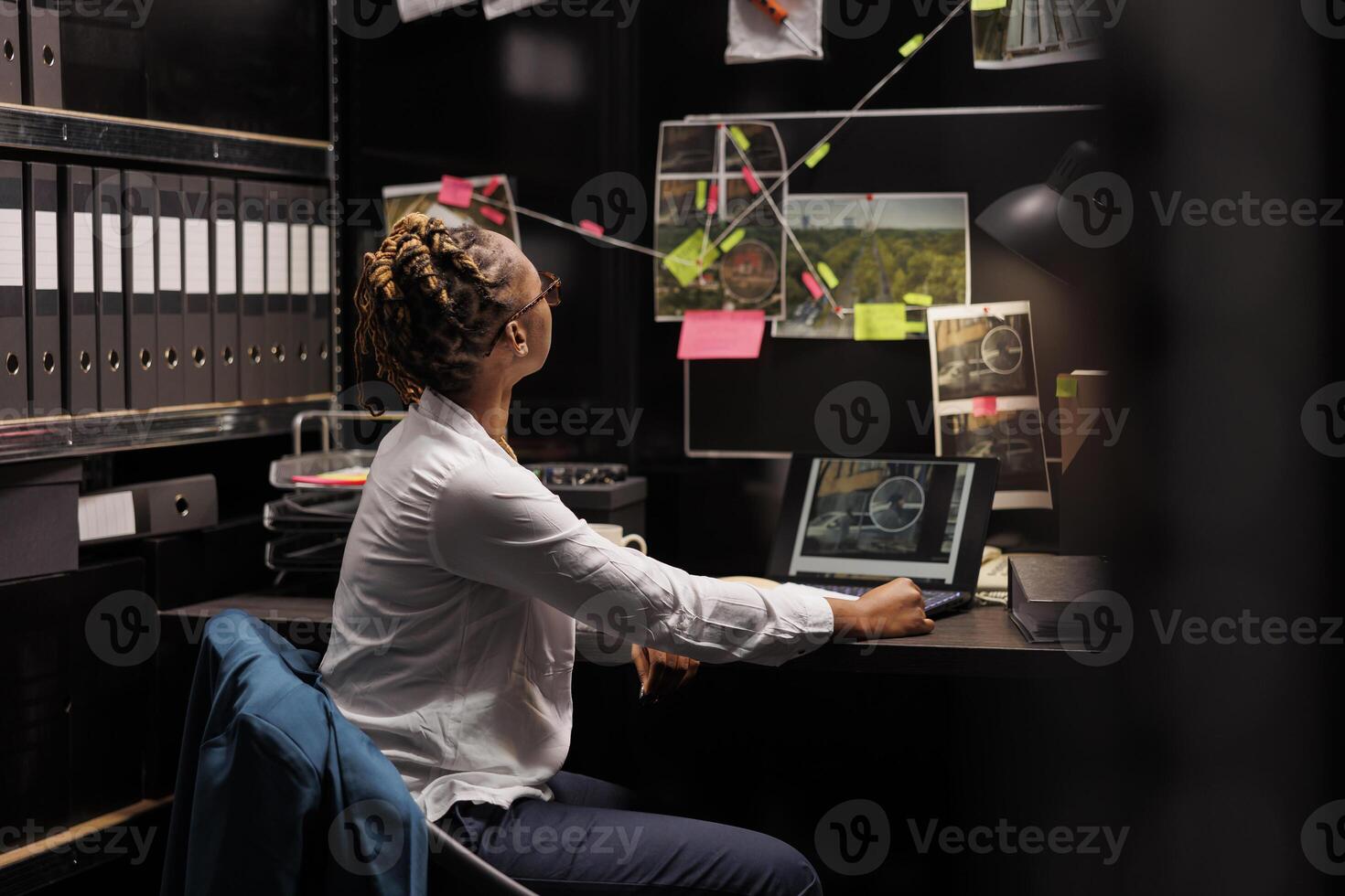 African american policewoman analyzing evidence board while solving crime. Law enforcement professional looking at clues connection scheme hanging on wall and searching insight photo