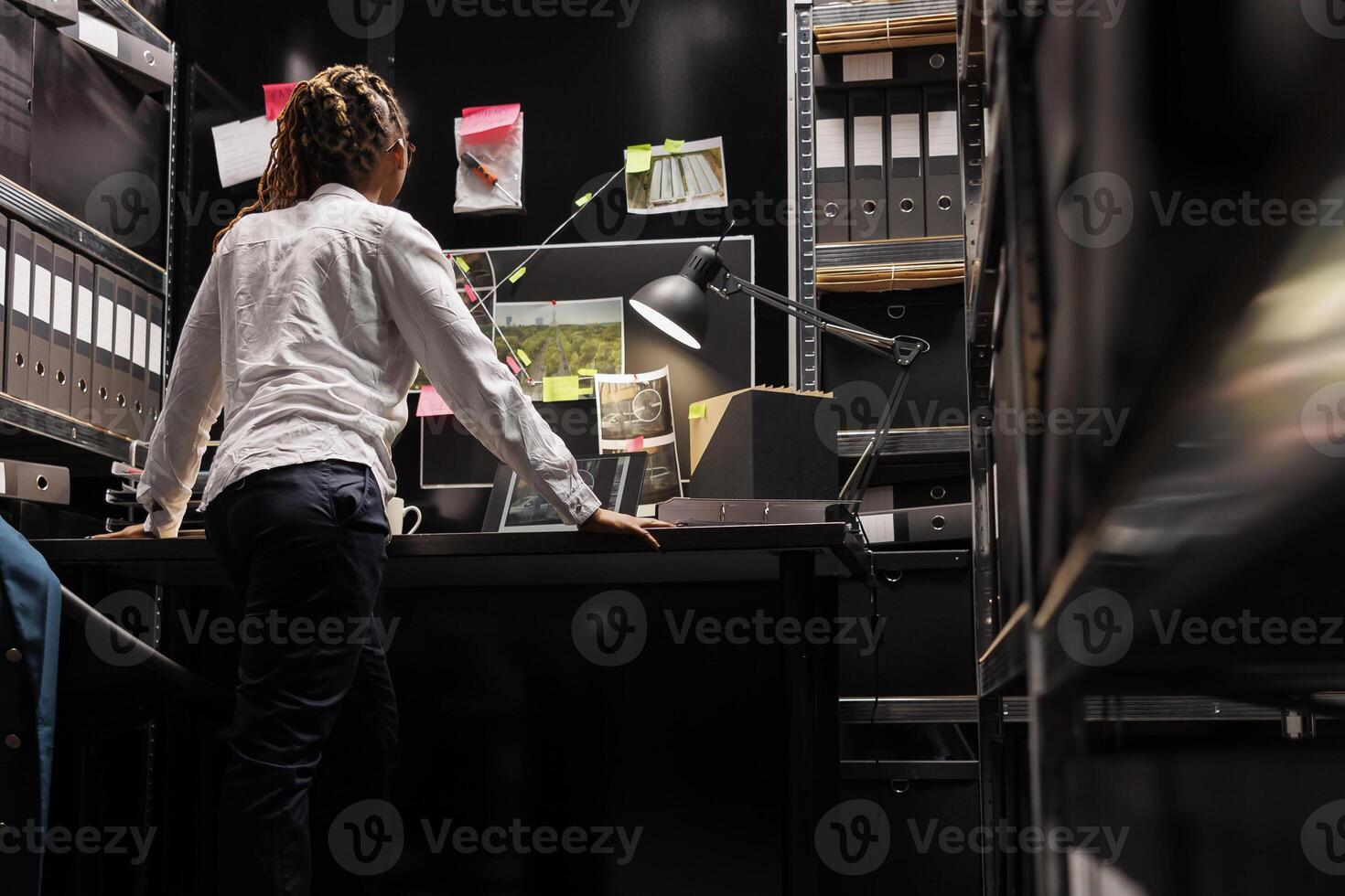 African american woman private detective looking at surveillance photos on laptop and analyzing clues. Investigator standing near desk, studying suspect photographs and evidence