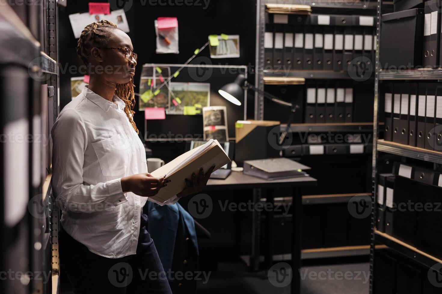 Law enforcement professional reading investigation materials in detective office room. African american woman investigator holding crime case file and looking at folders in shelf photo