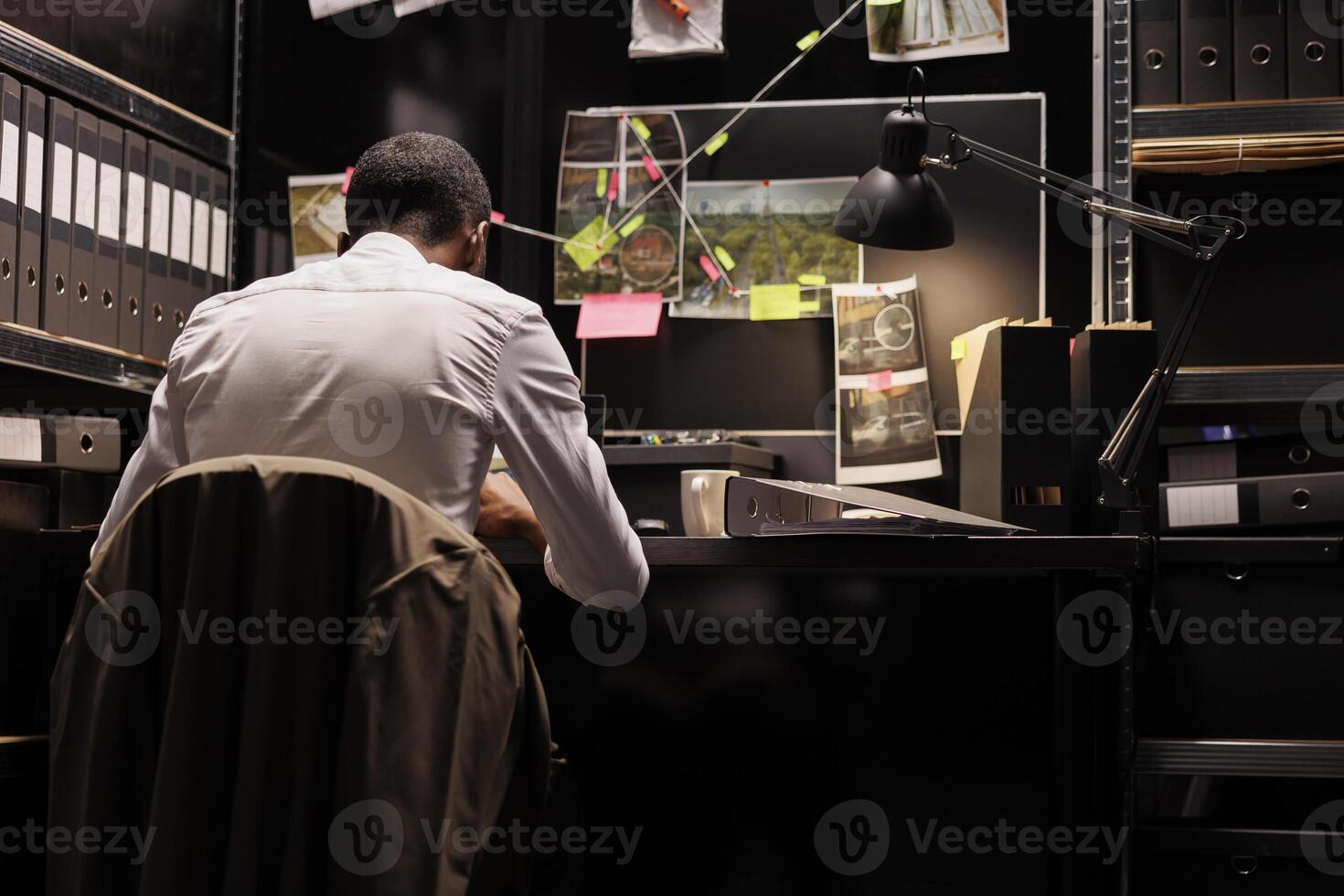 Detective sitting at workplace table and conducting investigation late. Private investigator studying clues and searching insight to solve complex crime case at night time photo