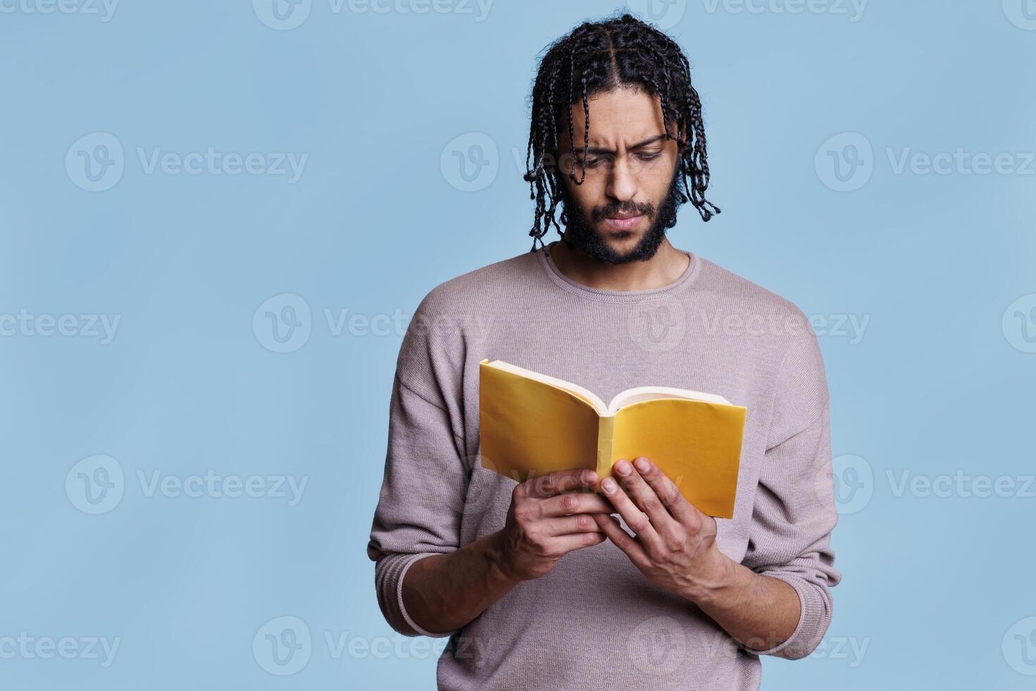 Concentrated arab man reading paperback book with yellow cover. Focused young person with thoughtful expression studying literature, holding softcover textbook on blue background photo