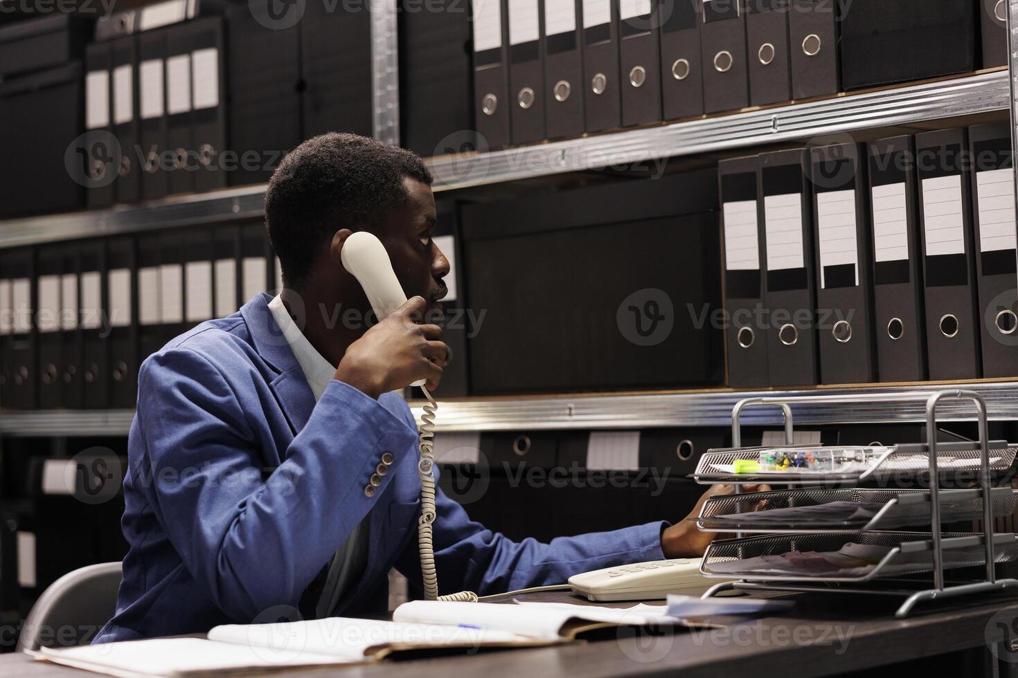 Police officer having remote conversation with inspector, discussing crime scene report using landline phone. African american detective working overhours at mysterious case in arhive room photo