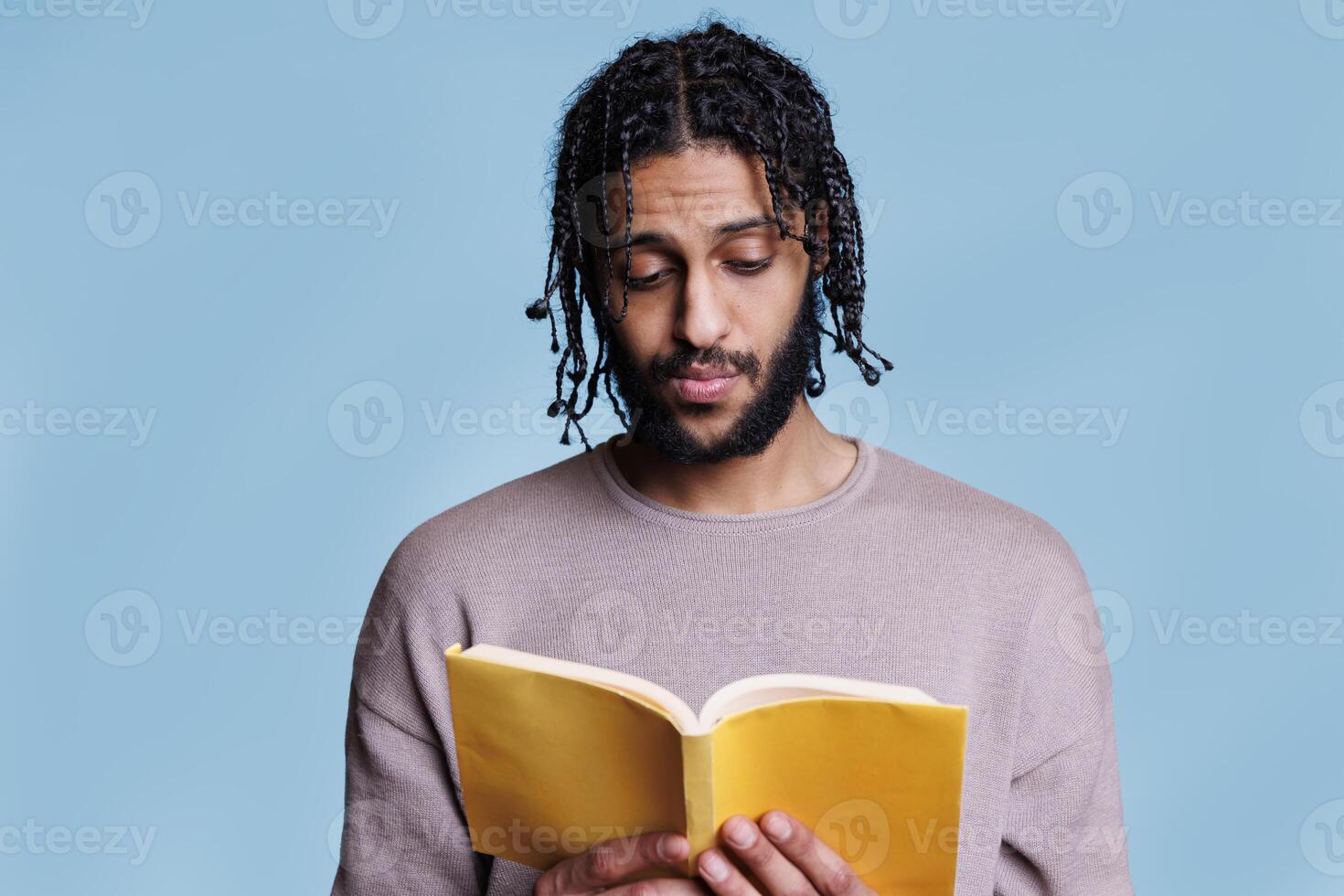 Serious concentrated arab man reading book and thinking about novel plot with pensive facial expression. Focused thoughtful young person holding paperback textbook with yellow cover photo