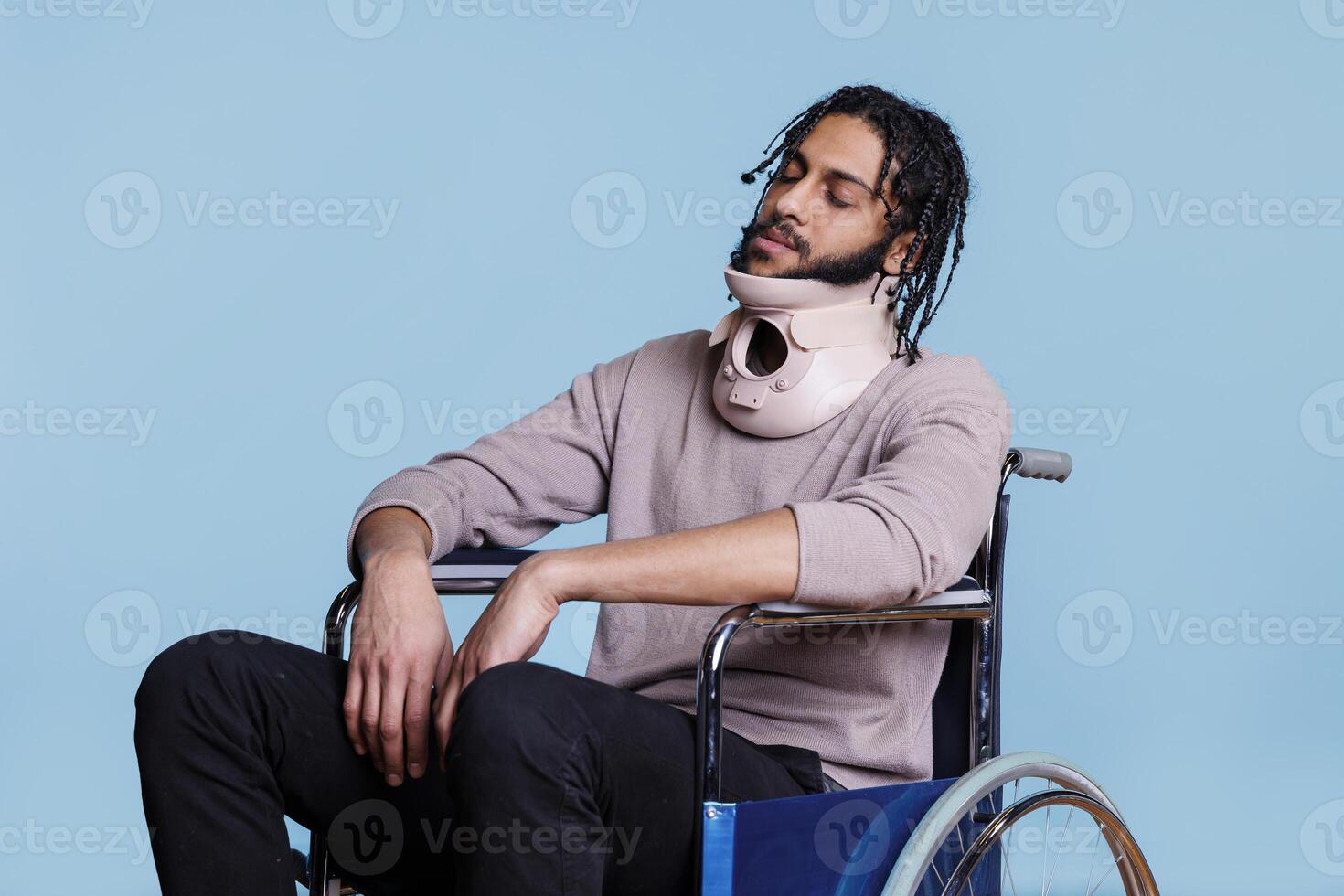 Arab man wearing cervical collar and sleeping while sitting in wheelchair. Young exhausted patient with closed eyes recovering from accident, using neck brace for spinal protection photo