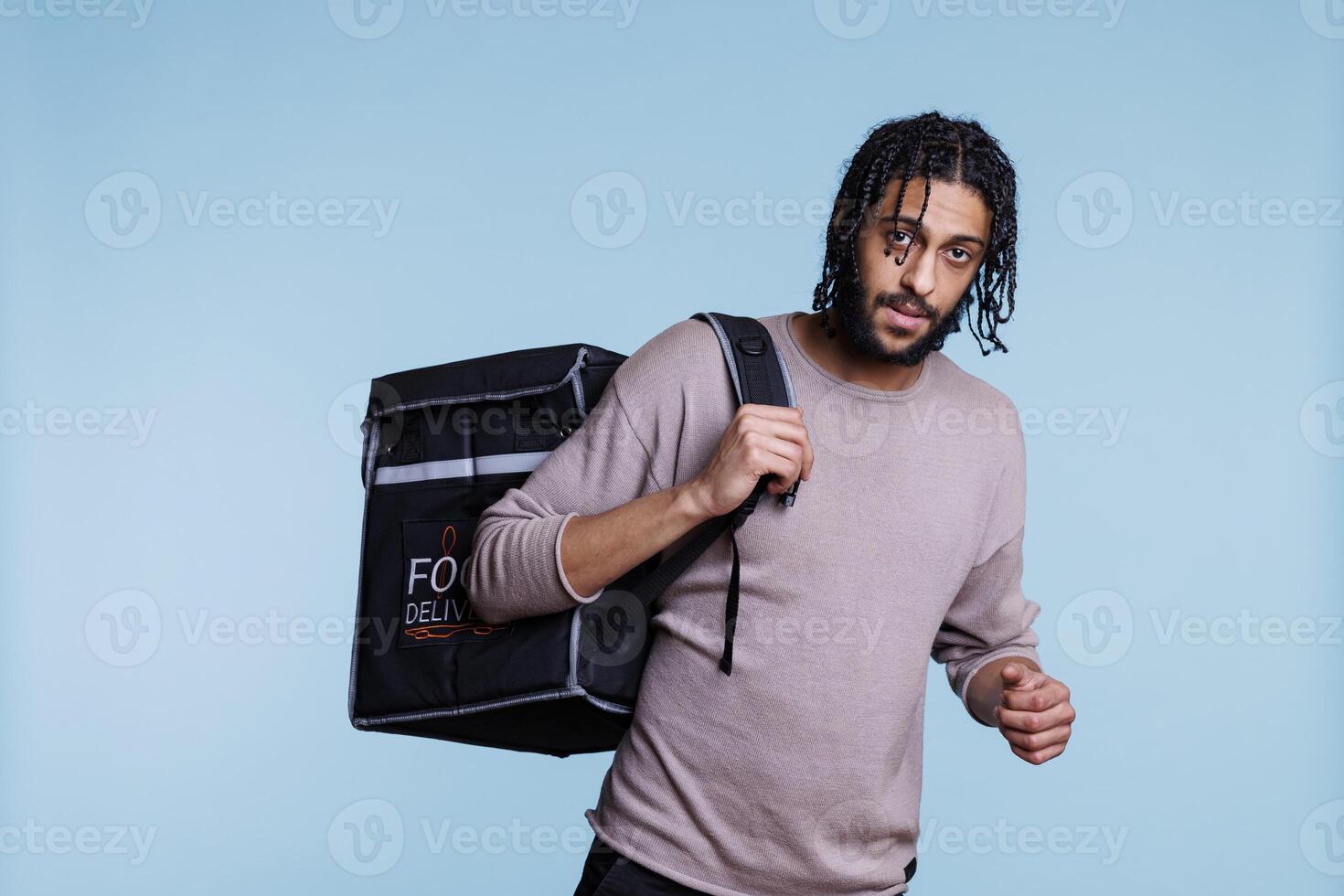 Food delivery service arab man carrying thermal bag portrait. Young deliveryman standing with backpack with restaurant take away meal and looking at camera on blue background photo