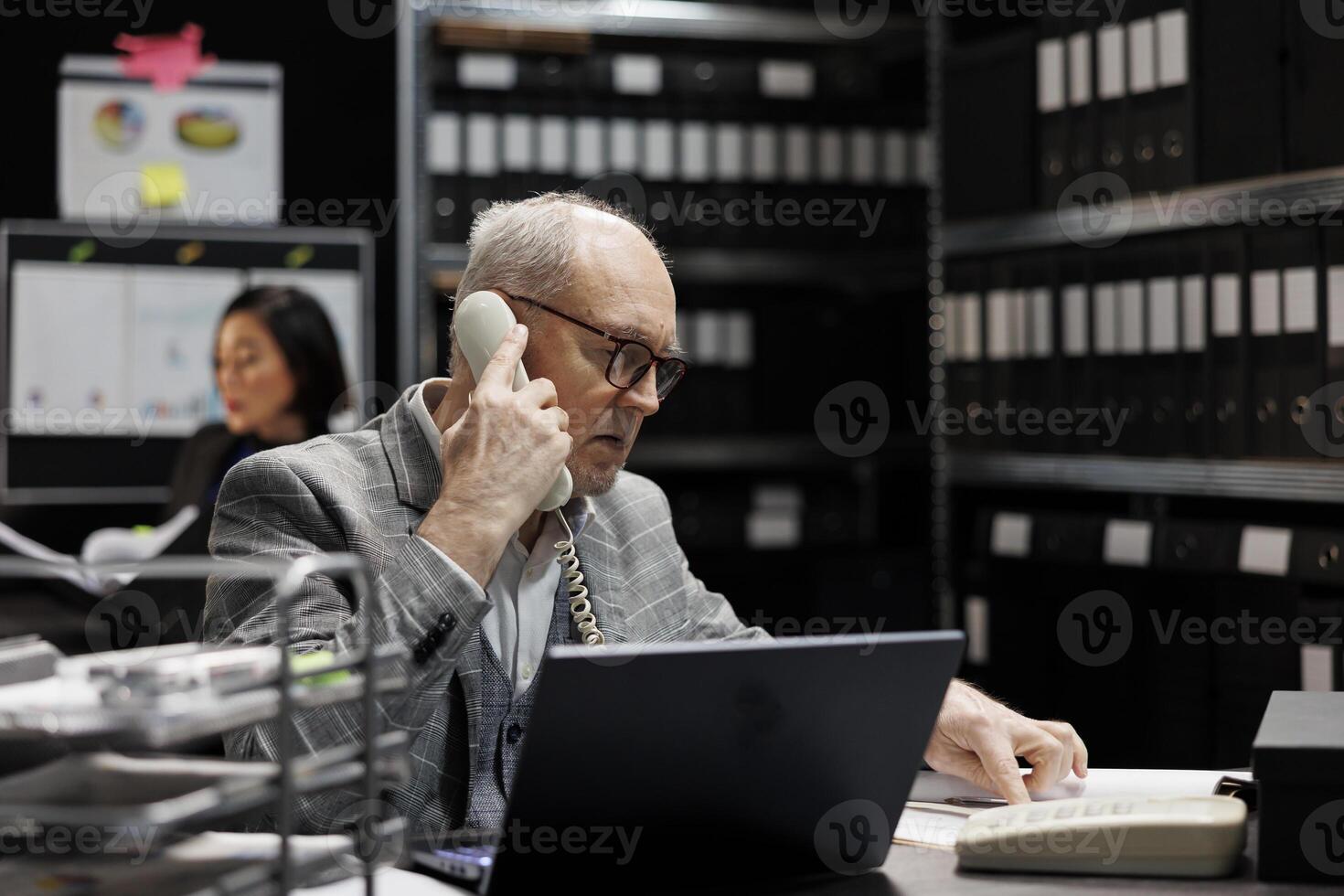 Accounting elderly businessman offering consultancy during phone call in bookkeeping workplace. Professional senior employee in archival depository filled with document folders and flowcharts photo