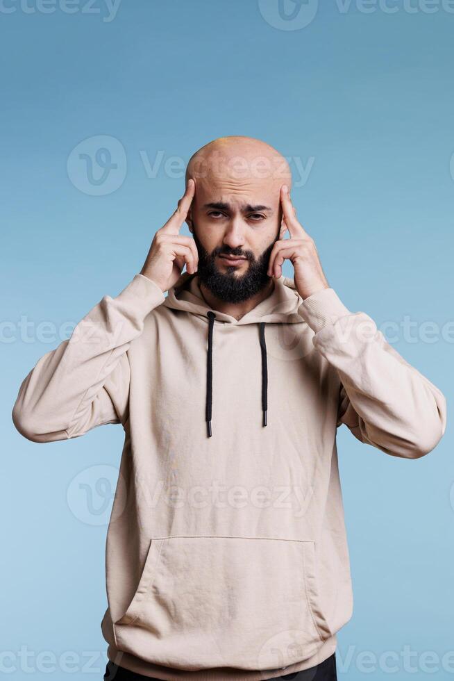 Arab man with migraine rubbing temples and looking at camera with pain grimace portrait. Young adult arabian bald bearded person touching head while suffering from headache photo