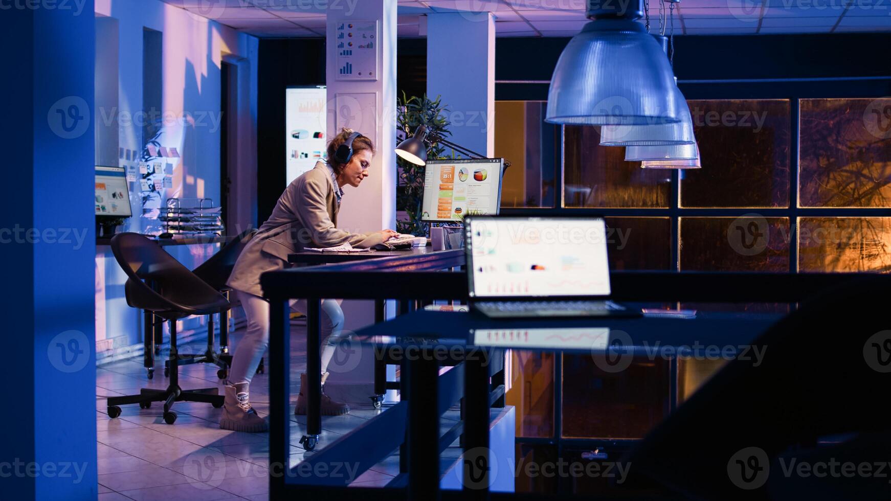 Happy employee analyzing graphs wearing headset, working at night to increase business profit. Advisor agent listening to music on headphones, checking startup analytics. Handheld shot. photo