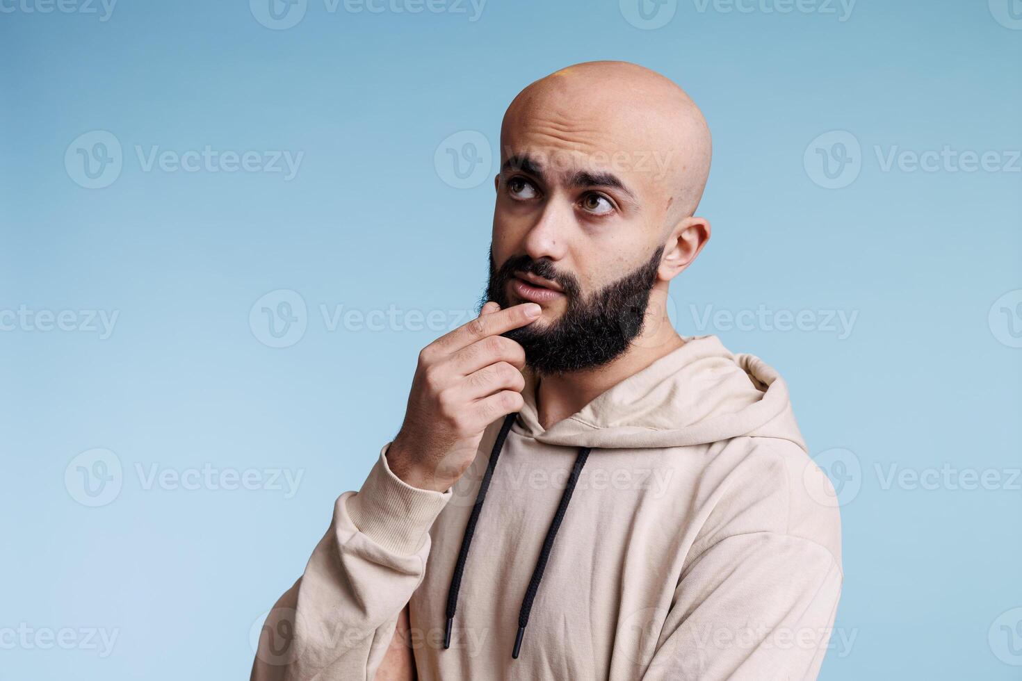 Arab man making decision, touching chin thoughtfully and looking away. Young bald bearded person with pensive facial expression contemplating, thinking about problem solving photo