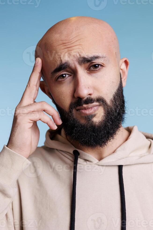 Tired arab man touching head while suffering from migraine studio portrait. Young bearded person feeling exhausted during headache and looking at camera on blue background photo