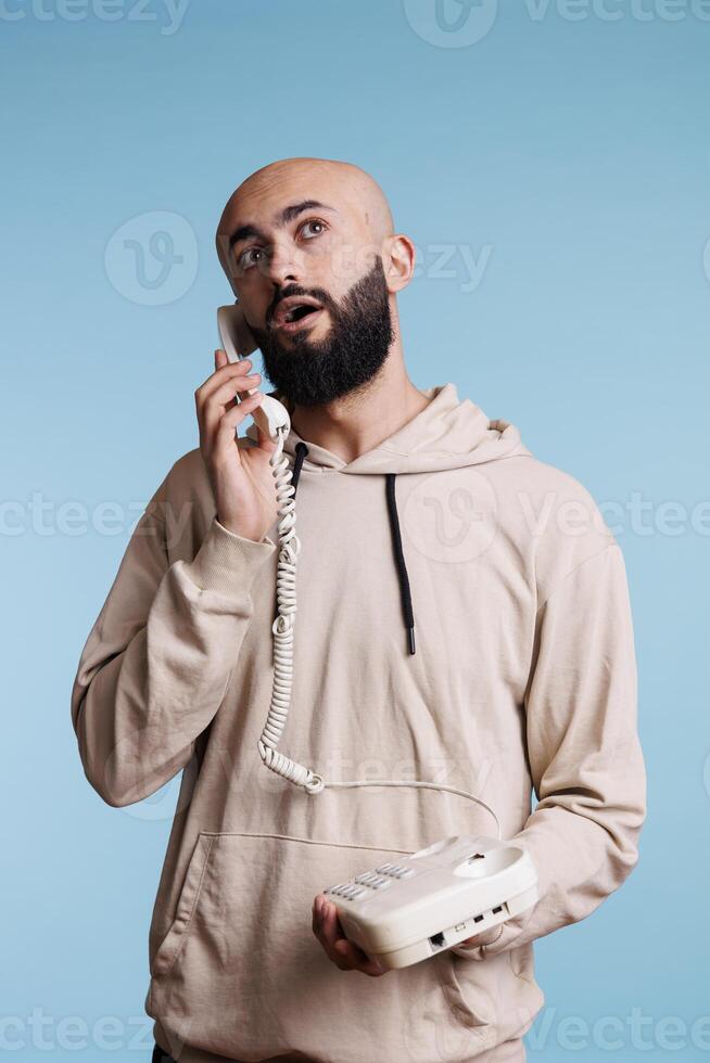 Thoughtful arab man speaking on corded landline phone and looking upwards while thinking. Young pensive person in contemplation holding retro telephone and having conversation photo