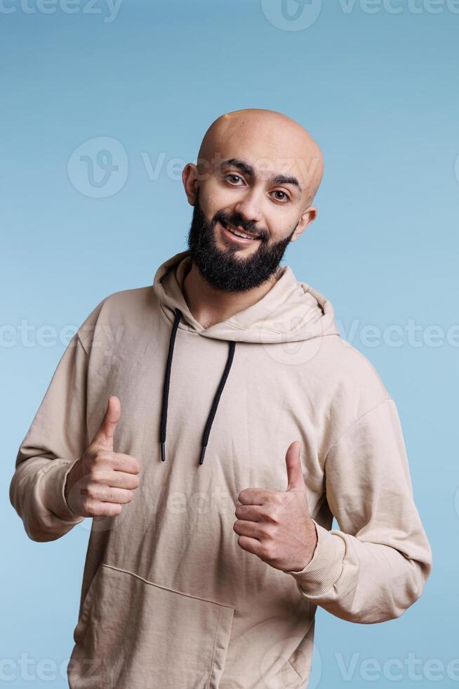 Cheerful arab man showing approval gesture with two thumbs up portrait. Smiling person with joyful facial expression standing with satisfaction sign while looking at camera photo
