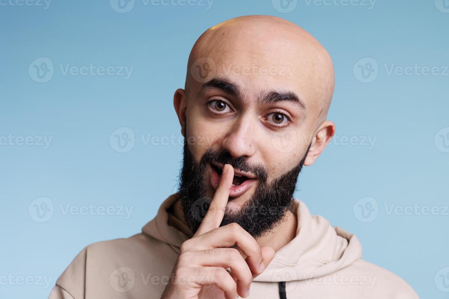 Young arab man making shh gesture with forefinger on lips and keeping silent portrait. Handsome bald bearded person showing quiet sign with finger on mouth and looking at camera photo
