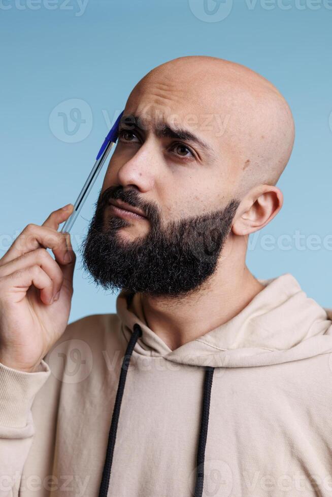 Young thoughtful arabian man rubbing head with pan while making decision and looking away. Puzzled person thinking, planning and dreaming with uncertain facial expression photo