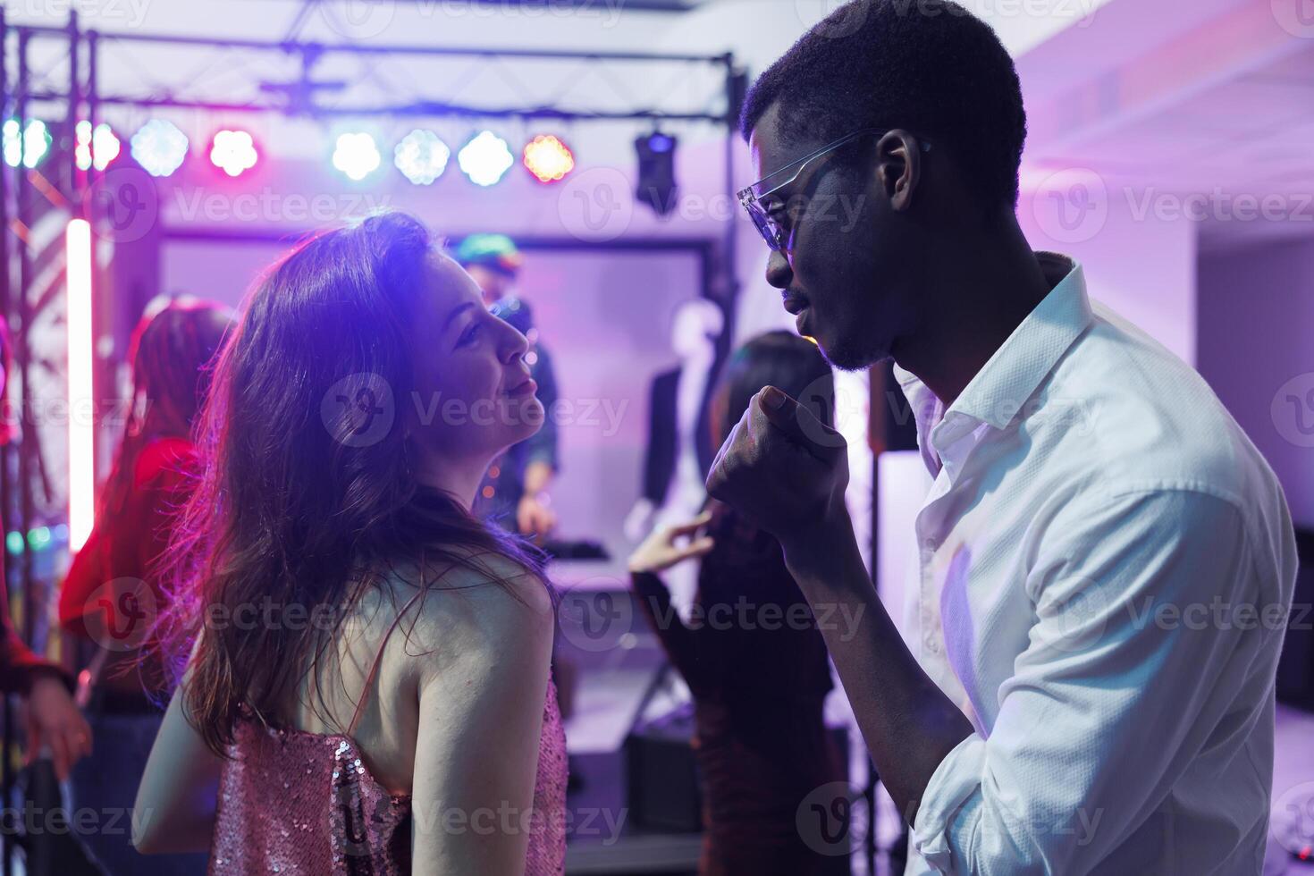 Couple in love dancing and clubbing while attending disco party in nightclub. Young diverse boyfriend and girfriend celebrating, moving to music rhythm on dancefloor illuminated with stage spotlights photo