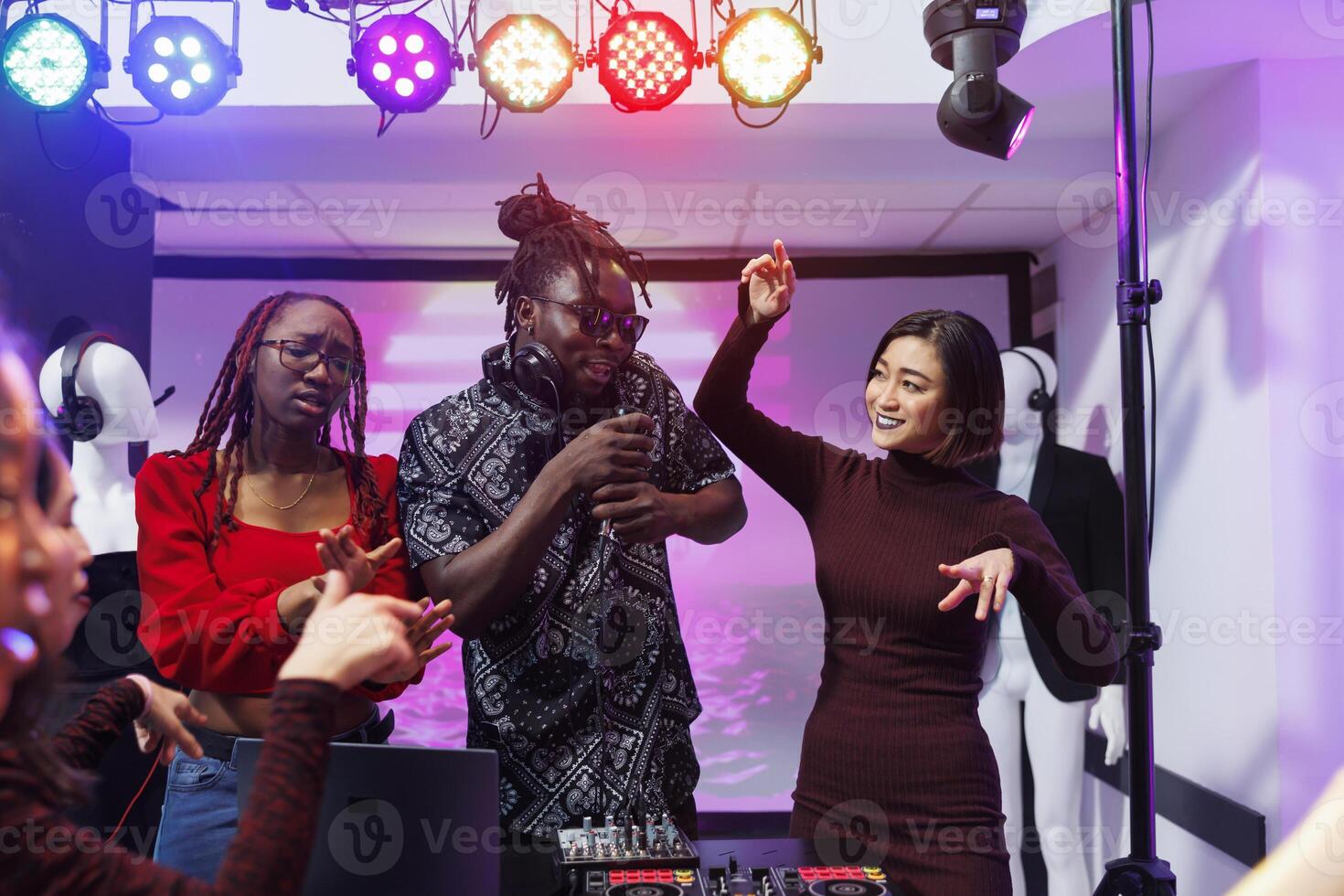 Band frontman singing and diverse women dancing on stage at discotheque in nightclub. Carefree musicians using dj station and microphone while performing at club disco party photo
