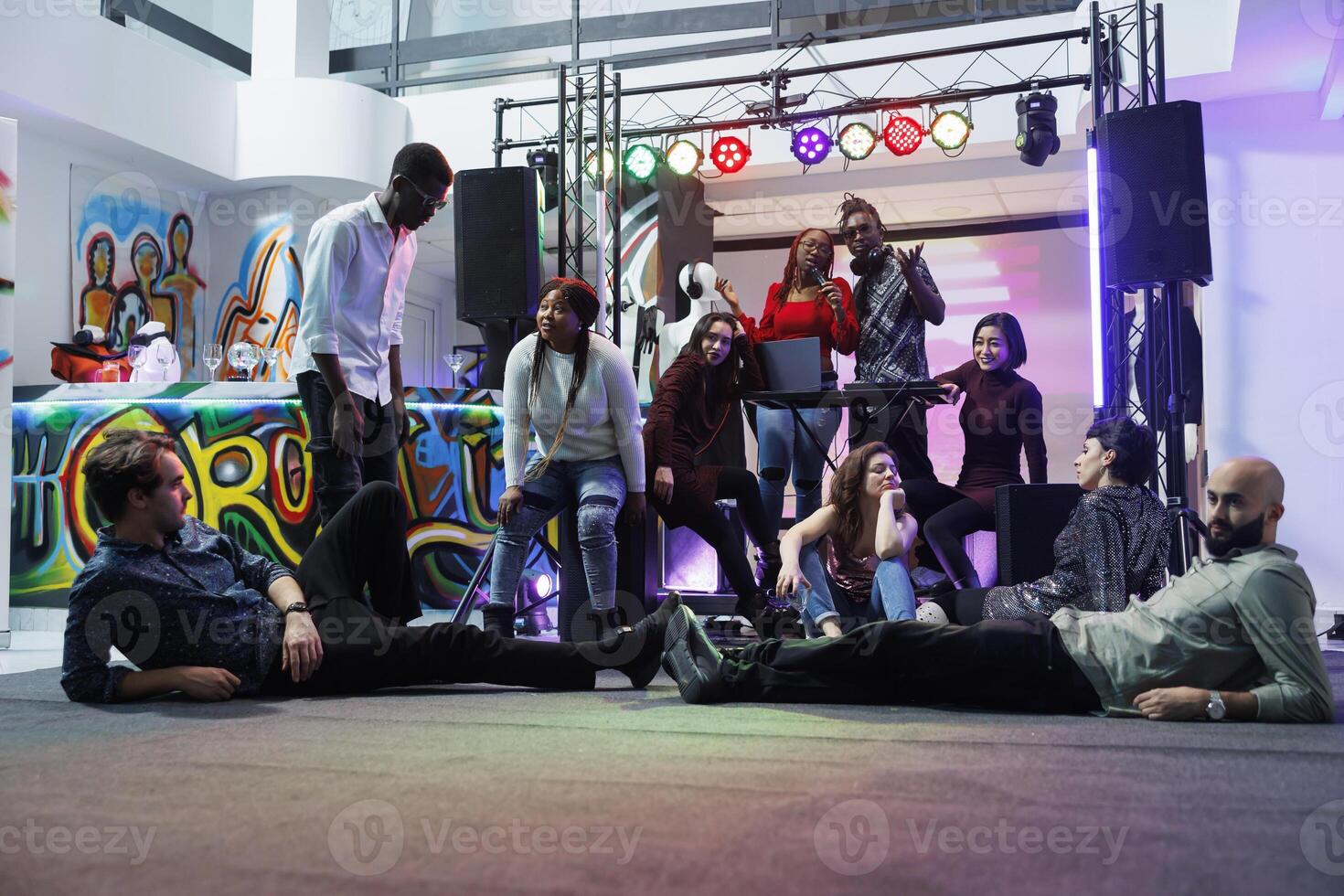 Music band performing on stage while diverse tired people lying and sitting on dancefloor. Exhausted young clubbers relaxing and listening to live music at club party gathering photo