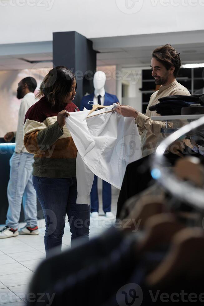 Fashion boutique diverse customer and assistant examining shirt fabric quality together. Clothing store caucasian man consultant providing woman client guidance on size and style photo
