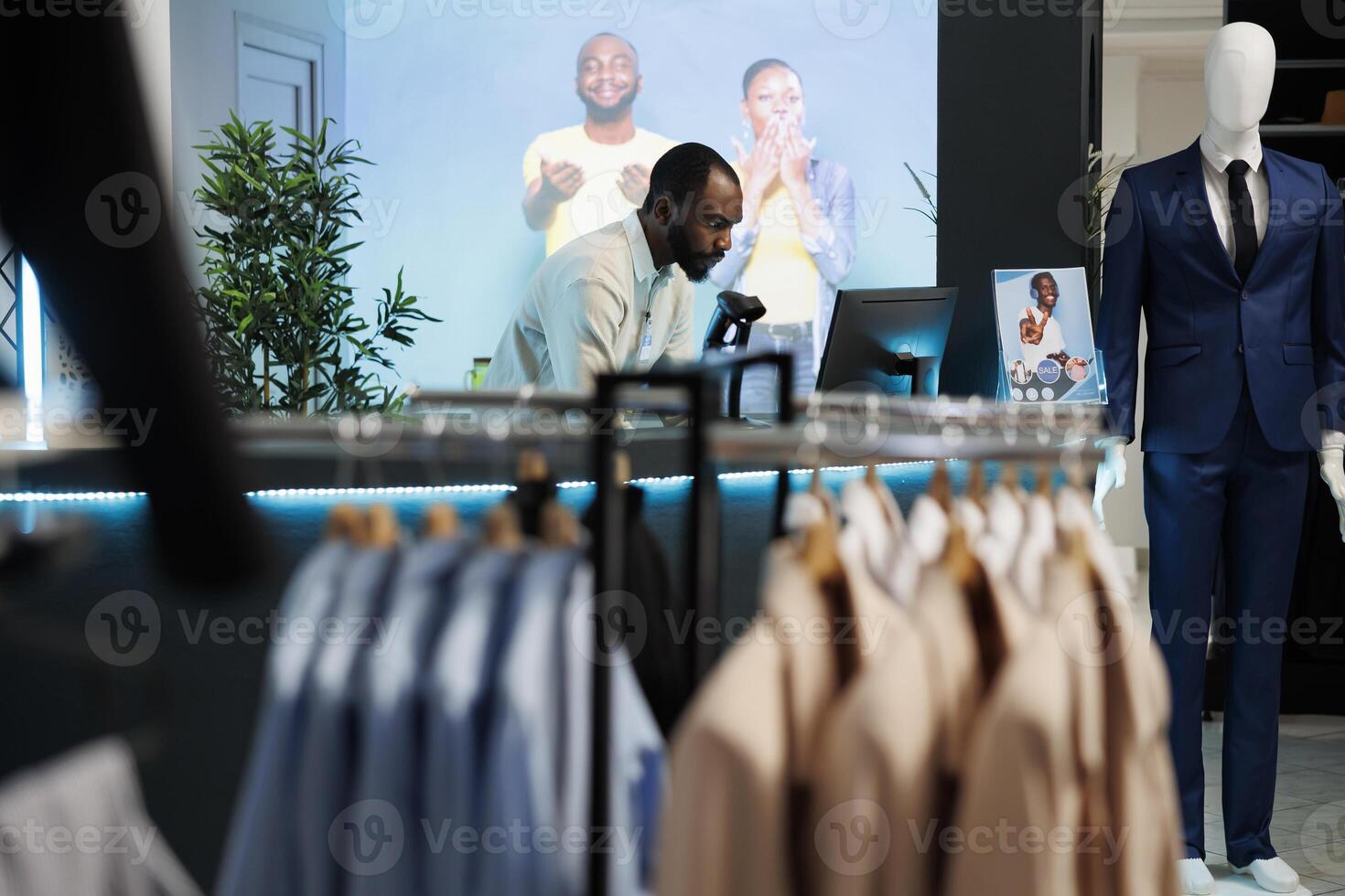 Man cashier working on computer at counter desk in trendy retail clothing store. Shopping mall boutique department african american assistant standing at checkout cash register photo