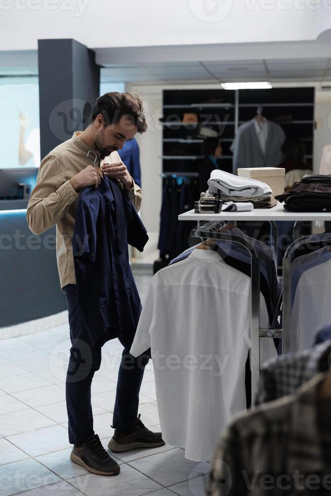 cliente participación camisa a cuerpo mientras elegir formal atuendo en Moda boutique ropa de caballero departamento. caucásico hombre molesto en de moda vestir y examinando estilo antes de comprando foto