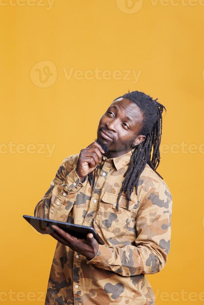 African american man browsing internet on digital tablet in studio over yellow background. Pensive young adult using wireless portable gadget surfing on social media app, watching video photo