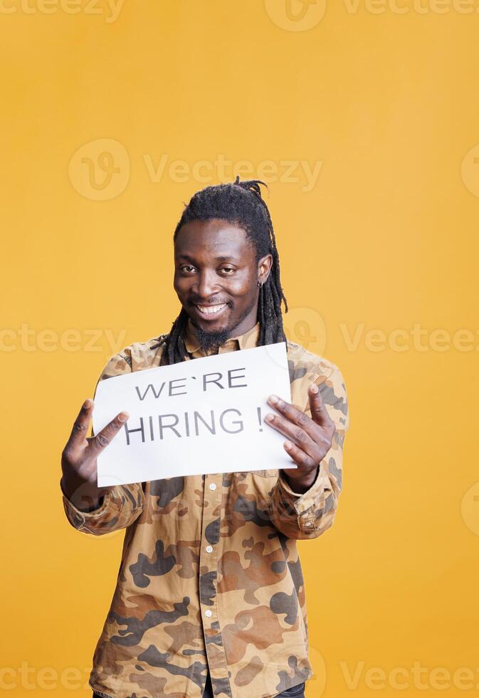 Company worker holding paper sign to make job offer in front of camera, hiring perfect candidate for career opportunity. African american employee asking people to attend job interview in studio photo