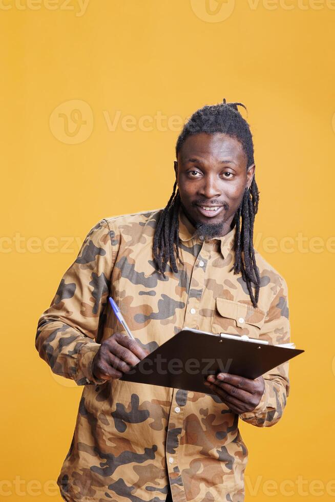 Confident person writing and taking notes on clipboard, checking papres with business job schedule in studio over yellow background. African american man looking at checklist, signing agreement files photo