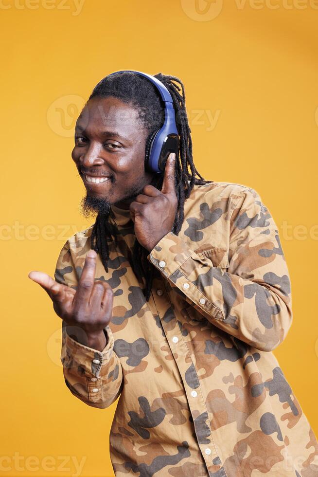 sonriente joven adulto disfrutando radio música en auriculares, en pie terminado azul antecedentes. africano americano hombre escuchando a mp3 canción, bailando y sensación contento utilizando inalámbrico auriculares foto