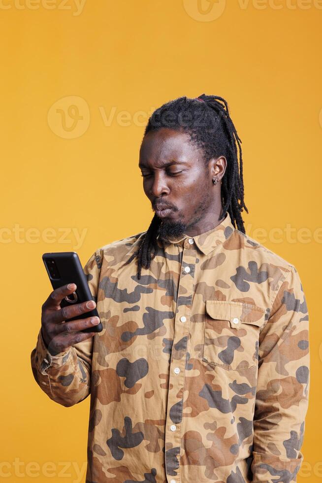 African american man reading good news on smartphone, scrolling through social media in studio over yellow background. Cheerful person searching information on phone, browsing on internet photo