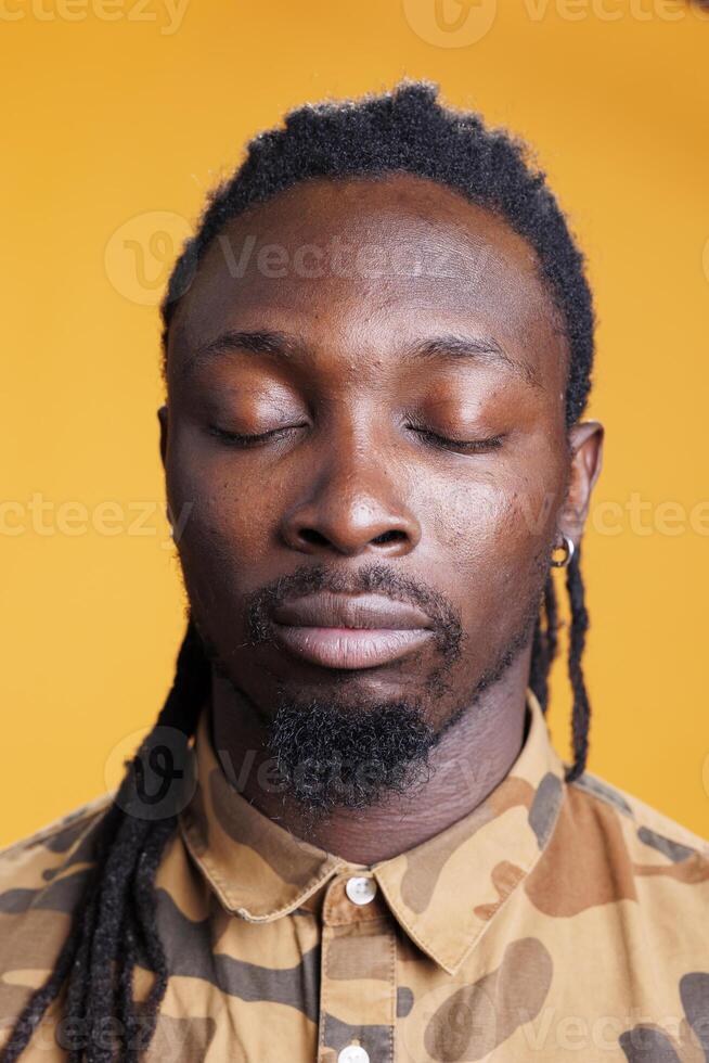 Relaxed man breathes deeply in studio, meditating with concentrated thoughts over yellow background. Concentrated african american person trying to relax, practicing wellness exercise. photo