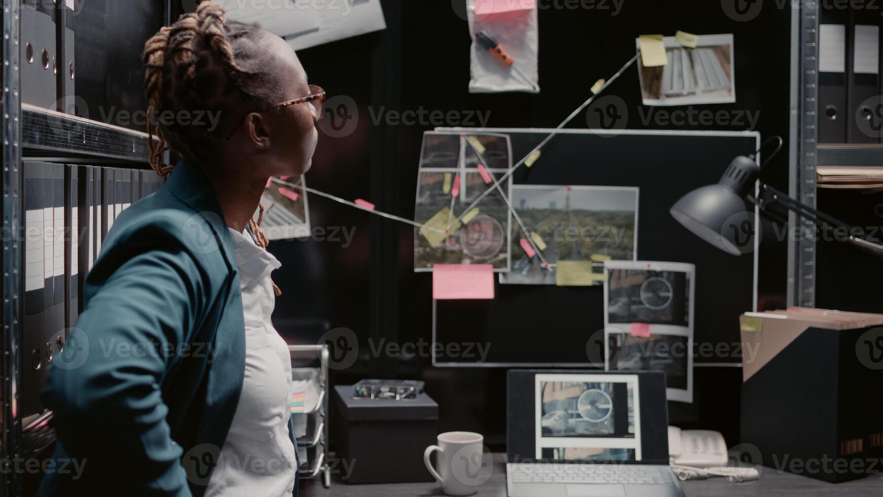 Professional officer reviewing surveillance photos on files to solve crime in police archive, reading witness reports. Young investigator analyzing forensic evidence attached on detective map.