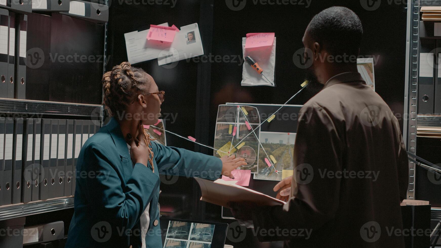 Detective partners analyzing criminal records in office, looking at archive photos. Police investigators examining evidence board and conducting investigation, pointing at clues. Handheld shot. photo