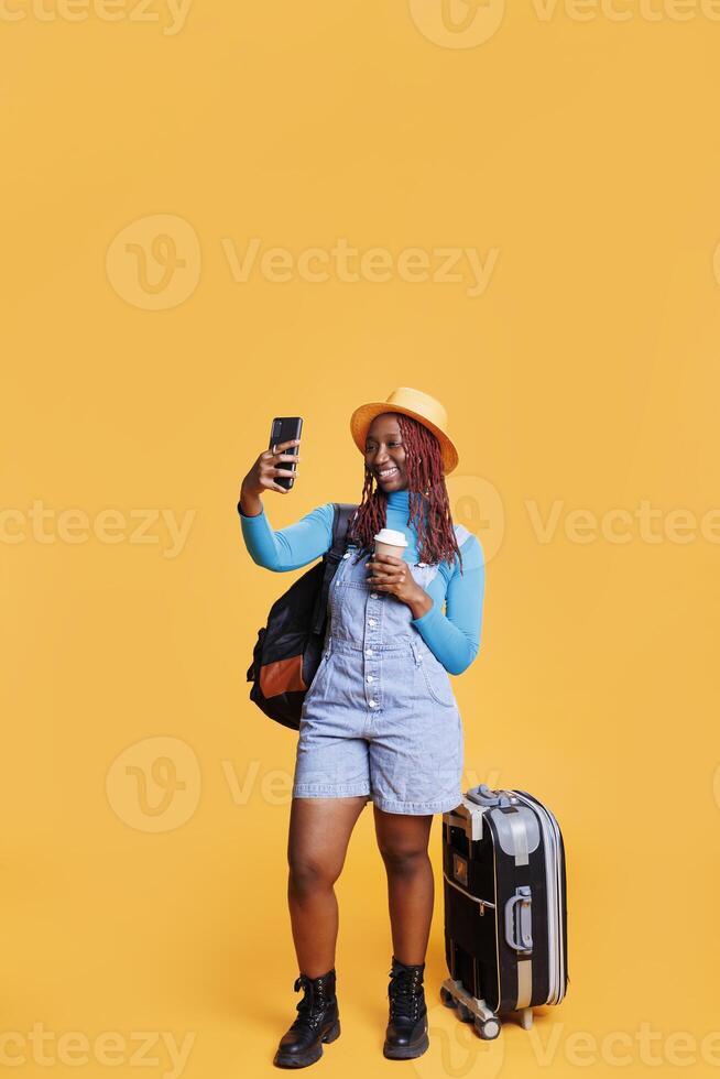 viajero tomando fotos en vacaciones viaje, haciendo tonto caras en teléfono inteligente y Bebiendo café. hembra persona teniendo divertido con imágenes en estudio, que lleva mochila y carretilla bolsas.