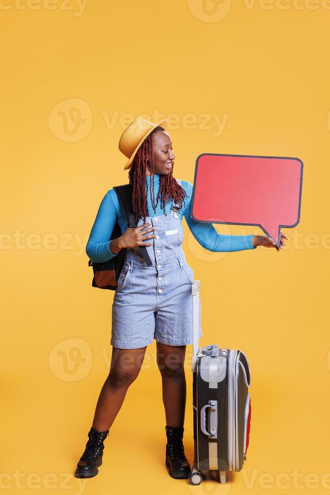 Smiling woman with empty speech bubble creating advertisement, leaving on vacation trip. Cheerful young adult showing isolated red billboard with copyspace in studio, cardboard. photo
