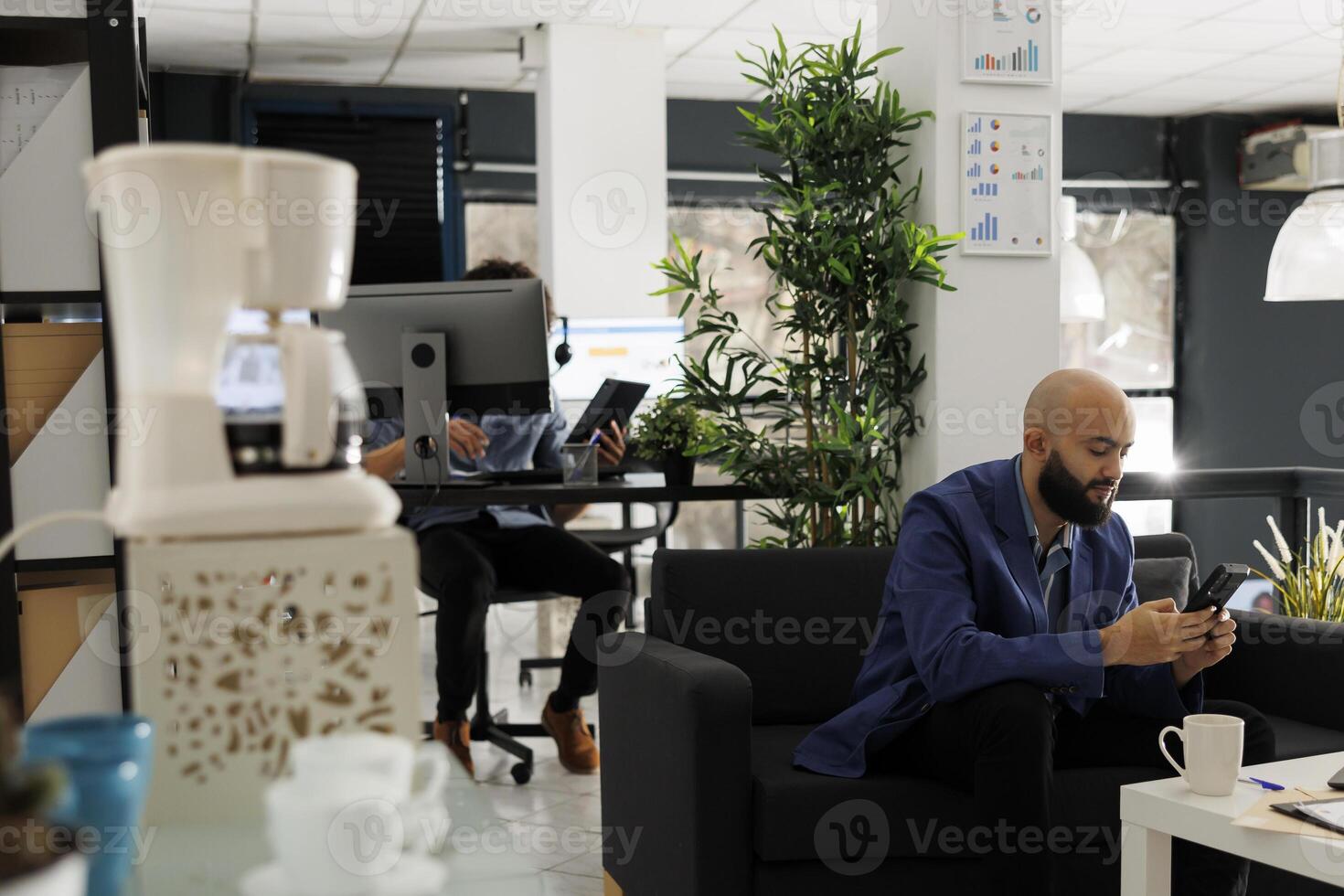 Arab business worker chatting with coworker using smartphone while working in modern open space. Marketing professional sitting on couch and texting in social media on mobile phone in start up office photo