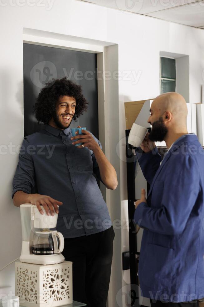 dos sonriente árabe empresarios teniendo café descanso y hablando en negocio oficina. empresa empleados en pie con té tazas mientras riendo y chateando en trabajo colaborativo espacio lugar de trabajo foto