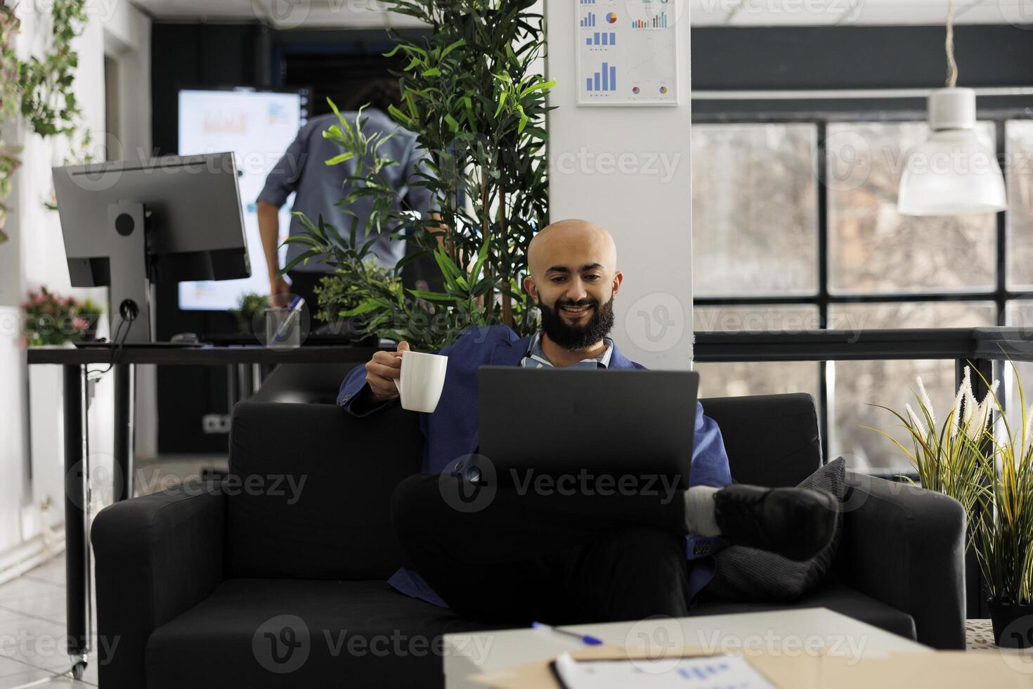 Smiling relaxed startup business employee drinking coffee and working on laptop in office. Happy arab man sitting on couch and holding tea mug while checking successful marketing results photo