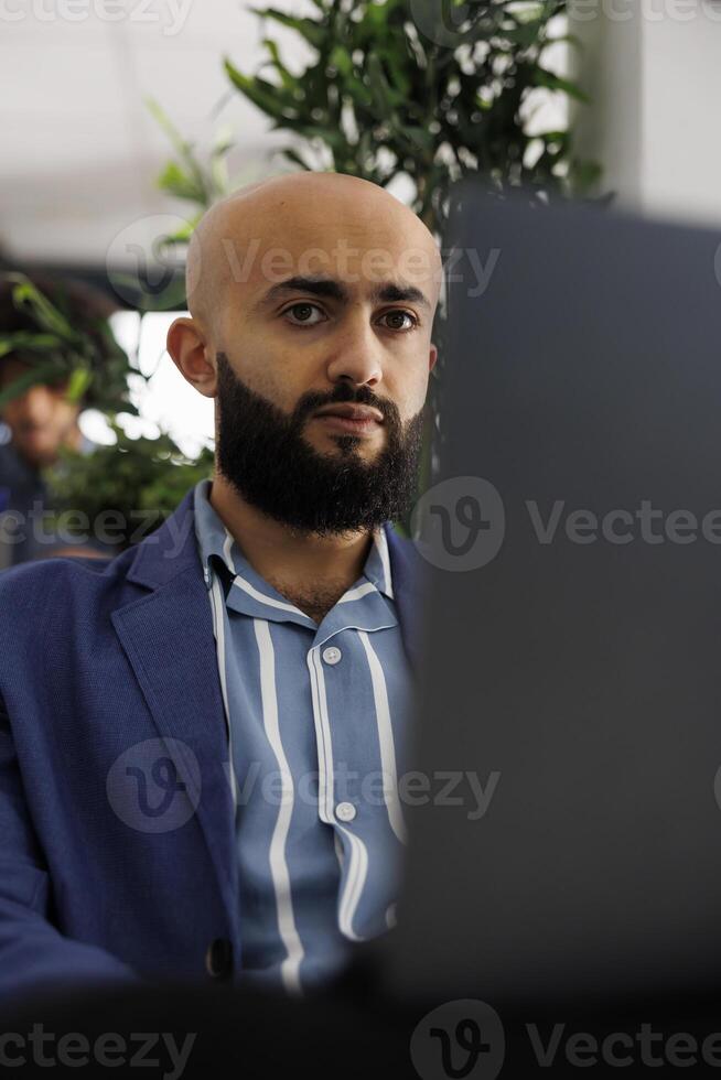 Arab businessman sitting in front of laptop at desk, preparing for video call with clients. Executive manager checking financial analytics report before presentation in coworking space photo